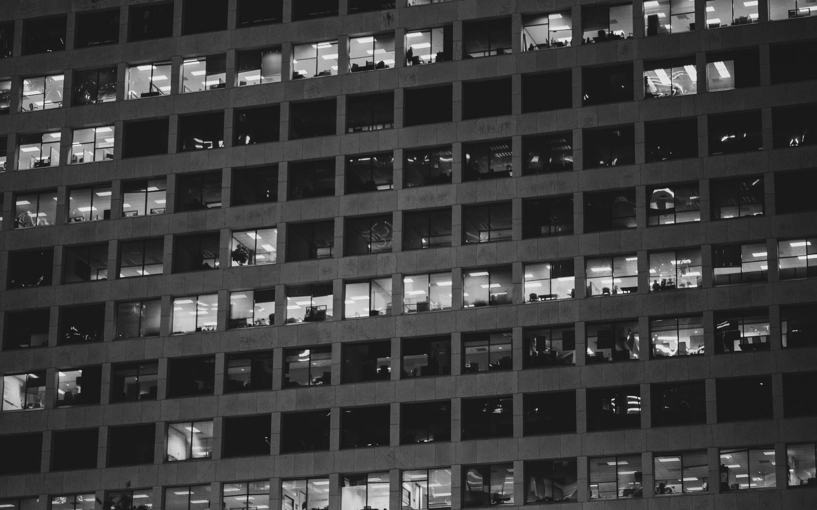 Une photo en noir et blanc montre l'extérieur d'un immeuble de bureaux la nuit, avec de nombreuses fenêtres éclairées et les silhouettes de personnes travaillant à l'intérieur — peut-être en train de discuter de l'alternance en entreprise alors qu'elles travaillent tard. La structure se compose de rangées et de colonnes de fenêtres identiques. "Alternance quels entreprises"