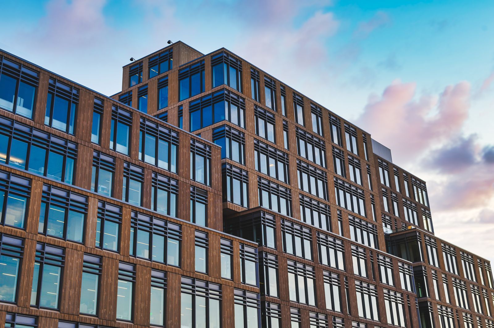 Un immeuble de bureaux moderne à plusieurs étages, doté de grandes baies vitrées et de touches de bois, se découpant sur un ciel bleu parsemé de nuages roses au coucher du soleil : un cadre idéal pour trouver une entreprise proposant une formation en alternance et favoriser votre évolution professionnelle.