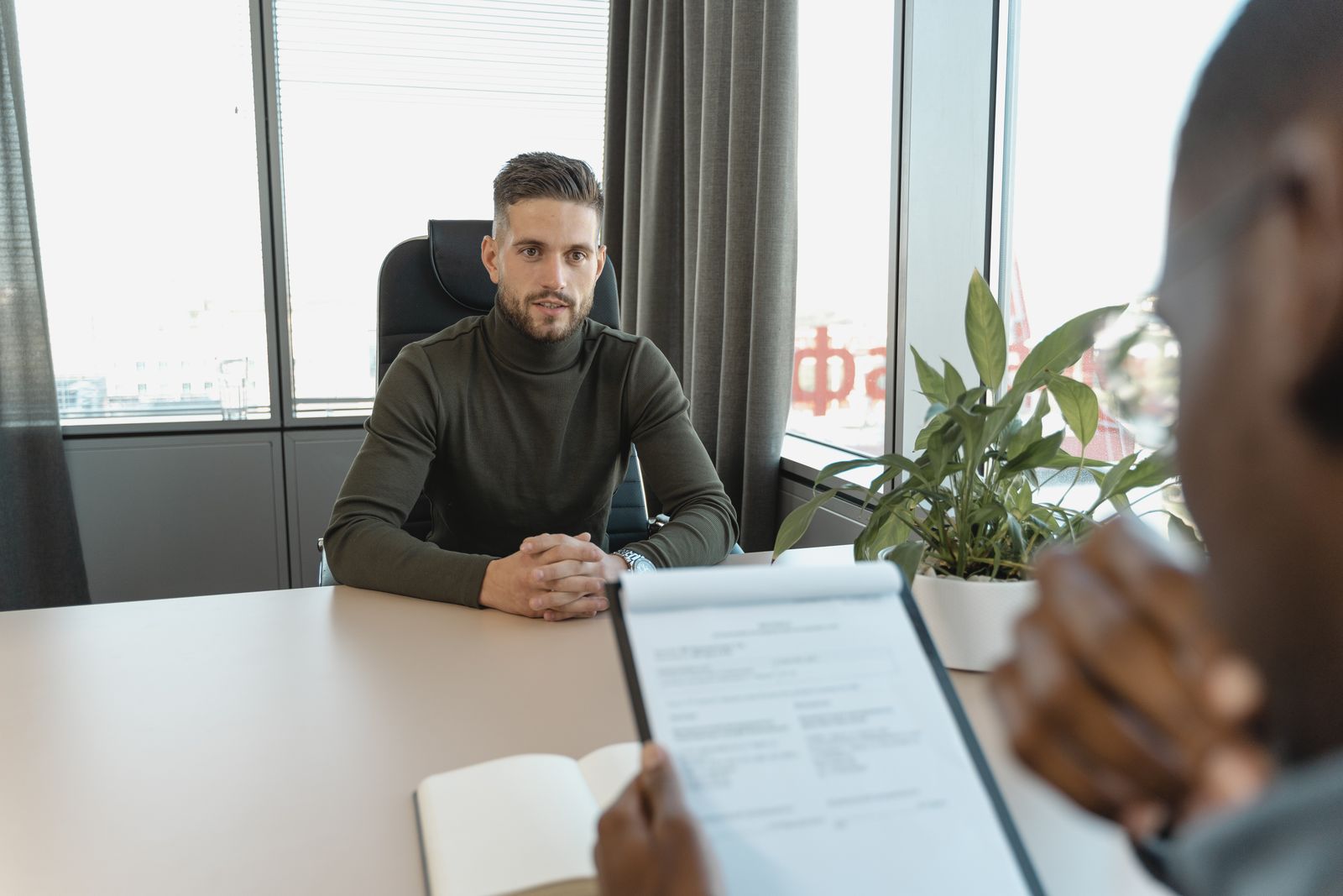 Un homme vêtu d'un col roulé vert est assis à un bureau, face à une autre personne tenant un bloc-notes et un CV, ce qui laisse supposer un entretien d'embauche dans un bureau moderne doté de grandes fenêtres et d'une plante en pot. Le recruteur demande ""pourquoi cette alternance ?"