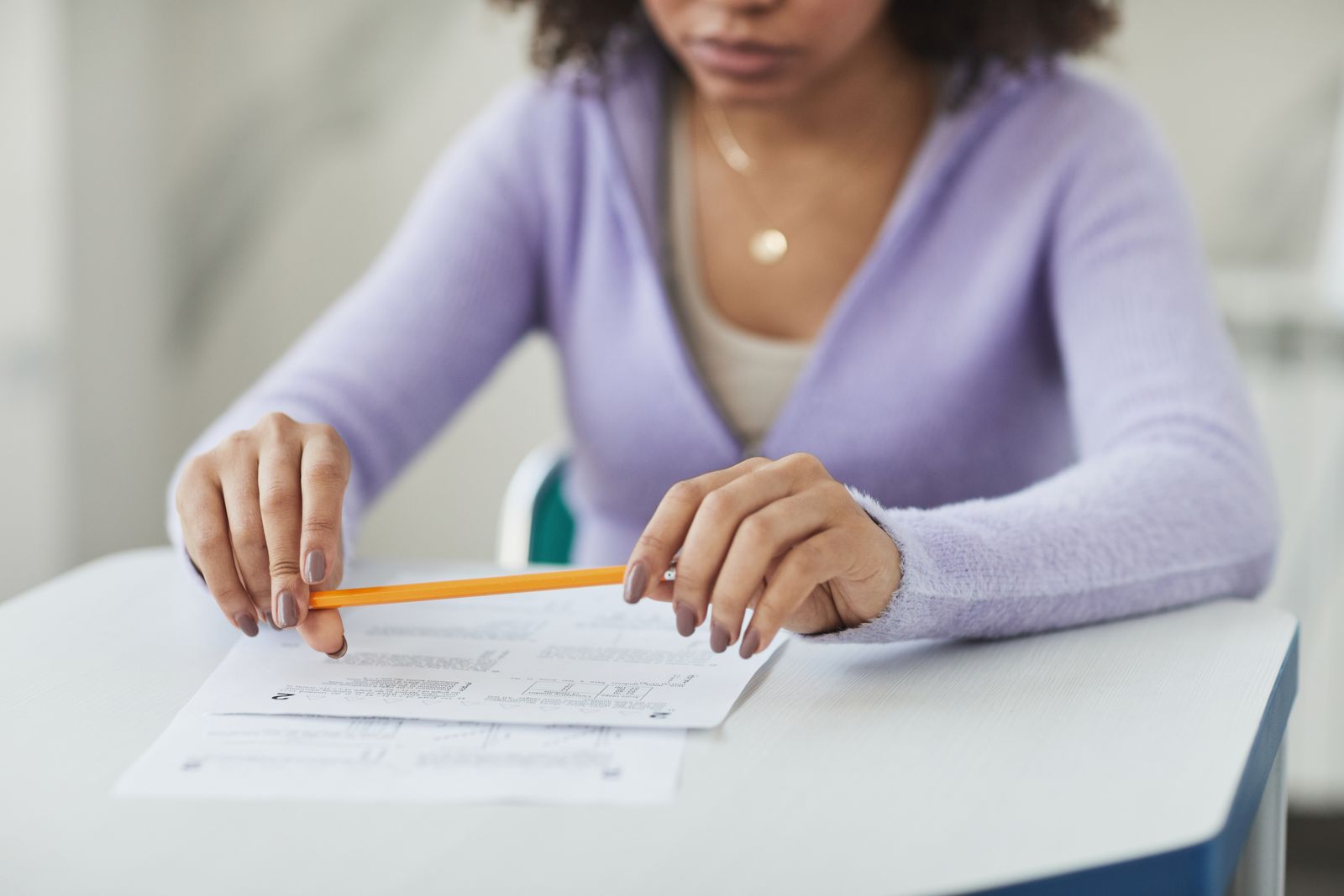 Une personne vêtue d'un pull violet clair est assise à un bureau, un crayon orange à la main, penchée sur un document comportant des graphiques et du texte ; elle semble concentrée et pensive — peut-être en train de relire les questions à préparer avant un entretien. Son visage est partiellement hors champ.