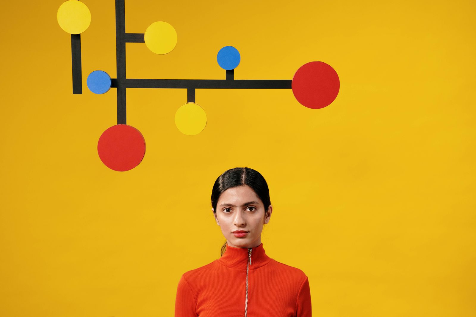 Une femme vêtue d'un haut orange à fermeture éclair se tient debout devant un fond jaune, incarnant la « mobilité interne », avec au-dessus d'elle une décoration murale géométrique composée de lignes noires et de cercles rouges, jaunes et bleus.