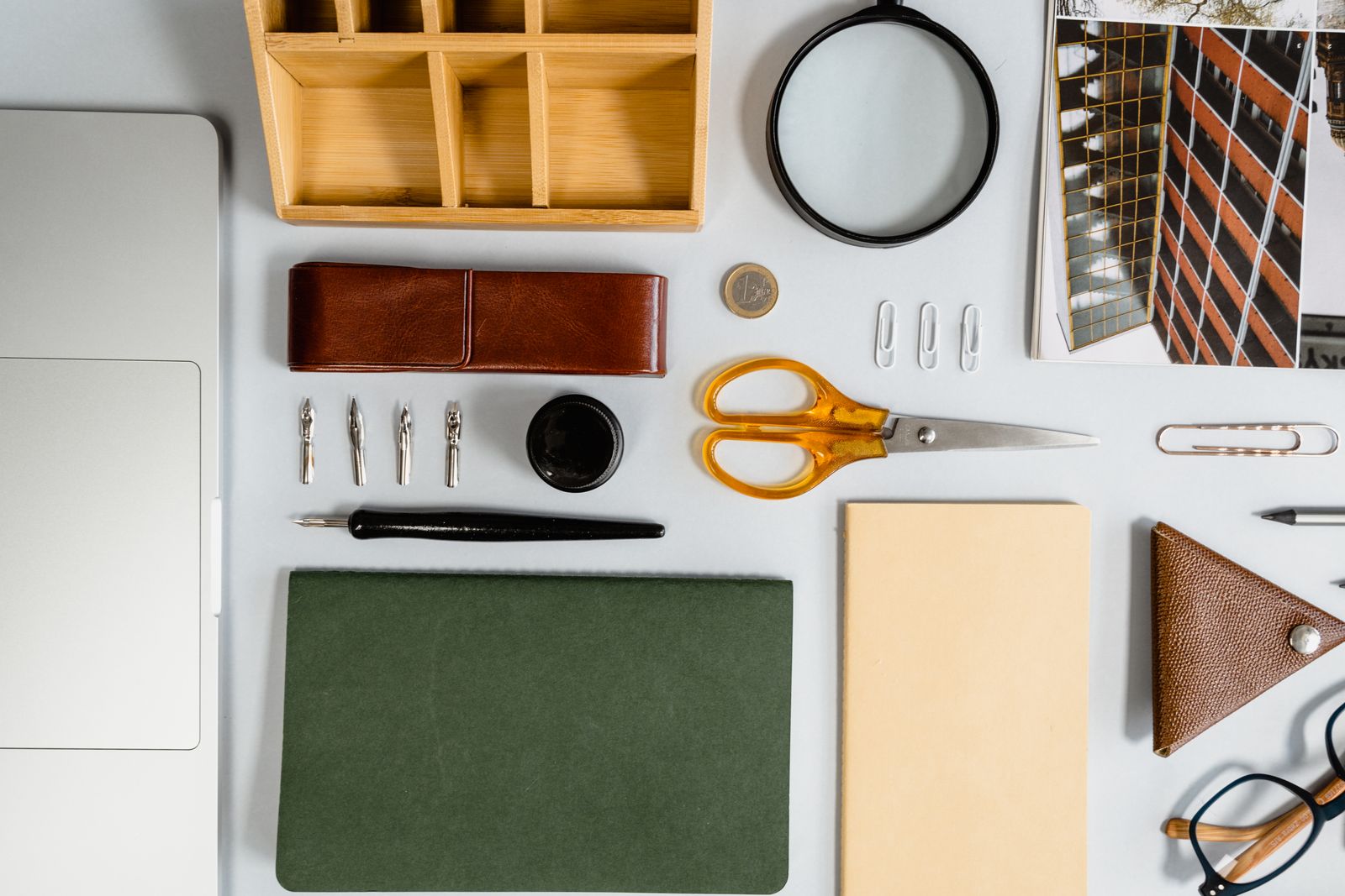 Une composition « flat lay » de fournitures de bureau, idéale pour organiser sa recherche d’emploi, comprenant des ciseaux, des stylos, des trombones, une loupe, des étuis en cuir, un cahier vert, un bloc-notes beige, des pièces de monnaie, un support pour ordinateur portable et des photos soigneusement disposées sur un fond clair.
