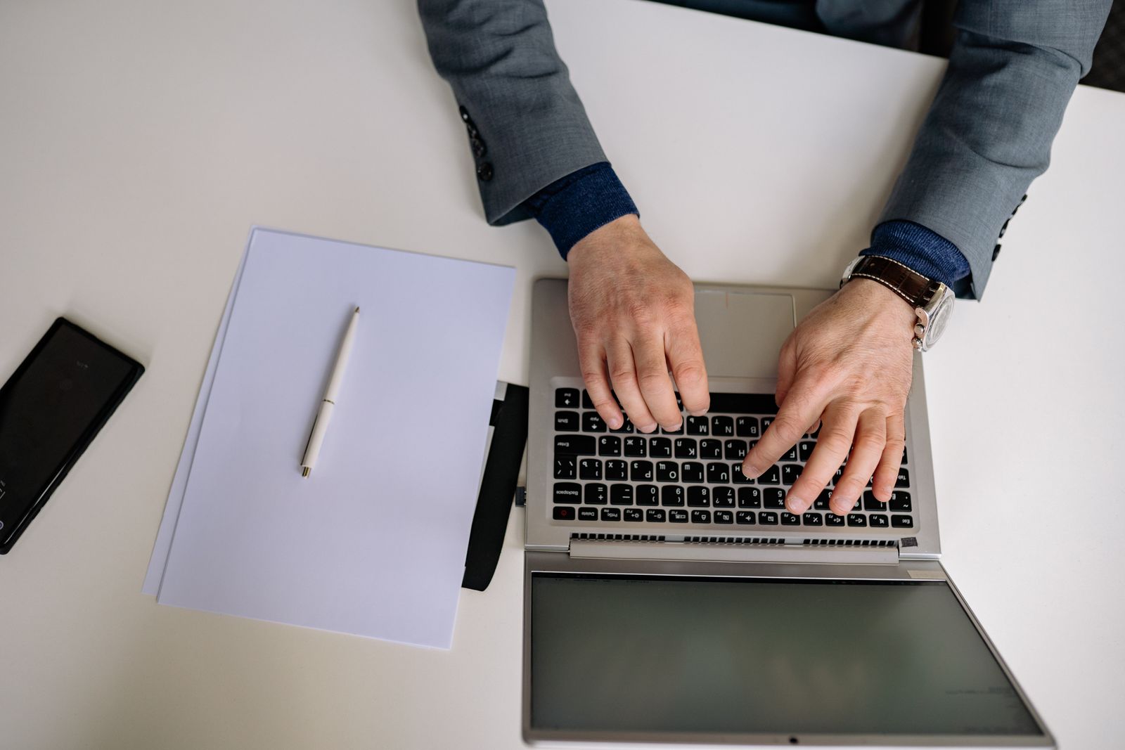 Une personne vêtue d'un costume gris tape sur un ordinateur portable posé sur un bureau blanc, s'apprêtant à faire un CV efficace. À proximité se trouvent une feuille de papier vierge, un stylo et un smartphone. La photo est prise en plongée.