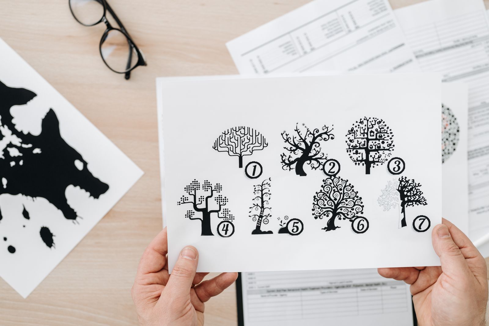 Une personne tient une feuille de papier sur laquelle figurent sept illustrations stylisées représentant des arbres et des cerveaux, chacune portant un numéro de 1 à 7, ce qui rappelle l'univers des tests de personnalité. Des feuilles, des verres et la silhouette noire d'un animal se trouvent sur le bureau à l'arrière-plan.