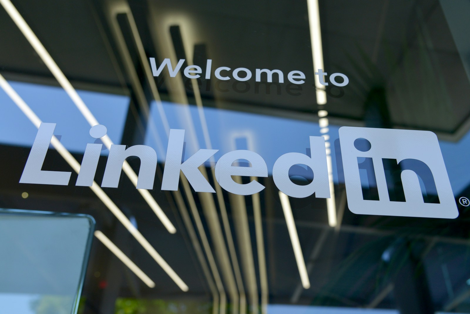 Le texte « Bienvenue sur LinkedIn », en lettres blanches, apparaît sur une porte ou une fenêtre vitrée, avec en arrière-plan un éclairage intérieur moderne et des reflets, soulignant ainsi comment vous pouvez utiliser LinkedIn pour trouver un emploi dans un cadre professionnel.