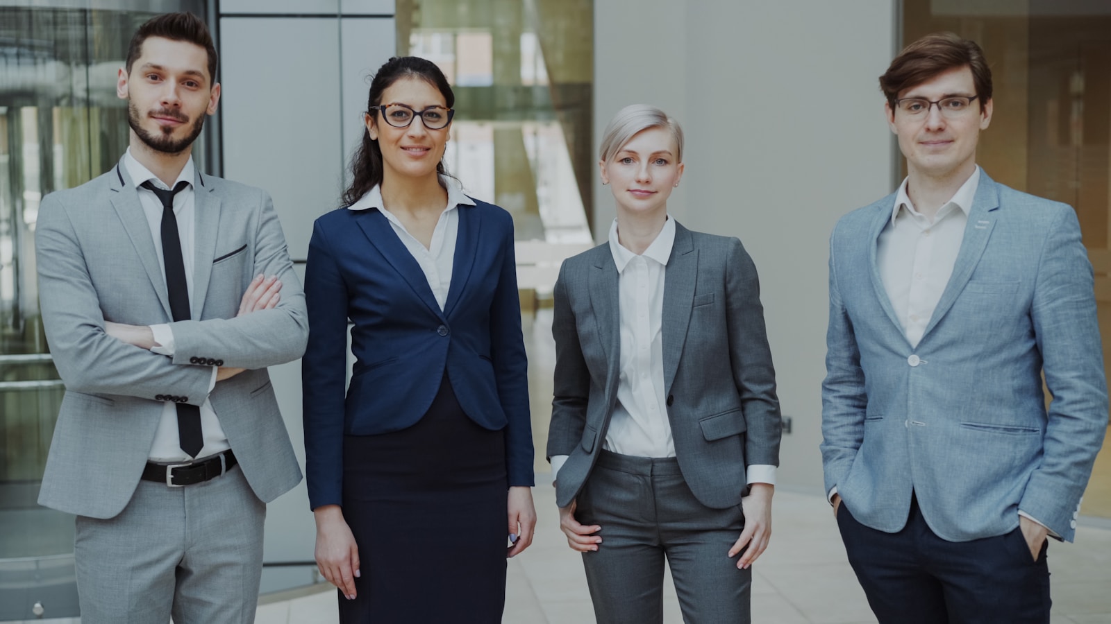 Quatre professionnels se tiennent à l'intérieur, vêtus de tenues professionnelles, affichant leur confiance dans leur projet professionnel. Trois d'entre eux portent des costumes gris et un autre un costume bleu marine ; tous sourient à la caméra, avec en arrière-plan un bureau au style moderne.
