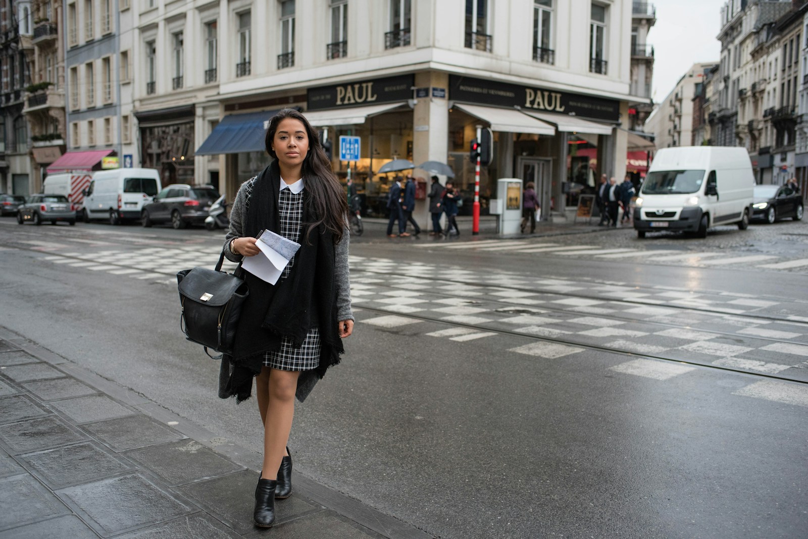 Une femme vêtue d'une robe à carreaux, d'un foulard noir et de bottines traverse une rue mouillée de la ville, peut-être en route pour retrouver un emploi, un grand sac noir et des papiers à la main. Derrière elle, on aperçoit une boulangerie PAUL ainsi que plusieurs piétons et véhicules.