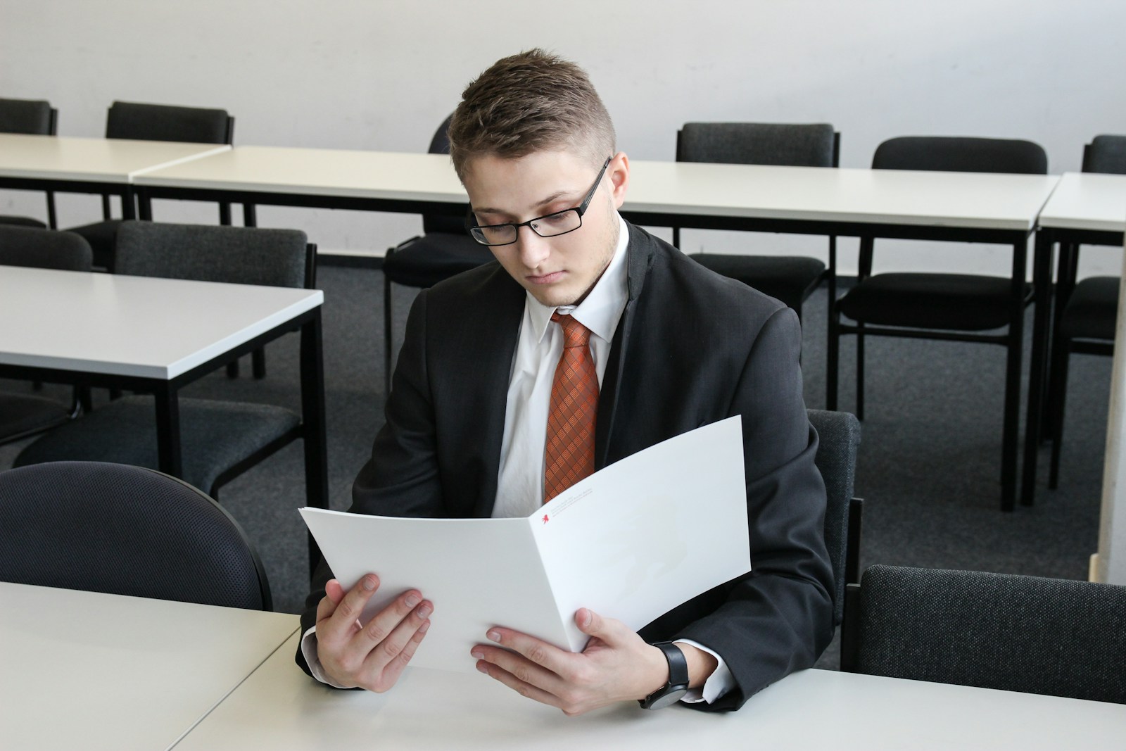 Un jeune homme en costume et cravate orange est assis à un bureau dans une salle de classe vide ; il consulte des documents contenus dans un dossier, sans doute pour préparer son CV stage étudiant.