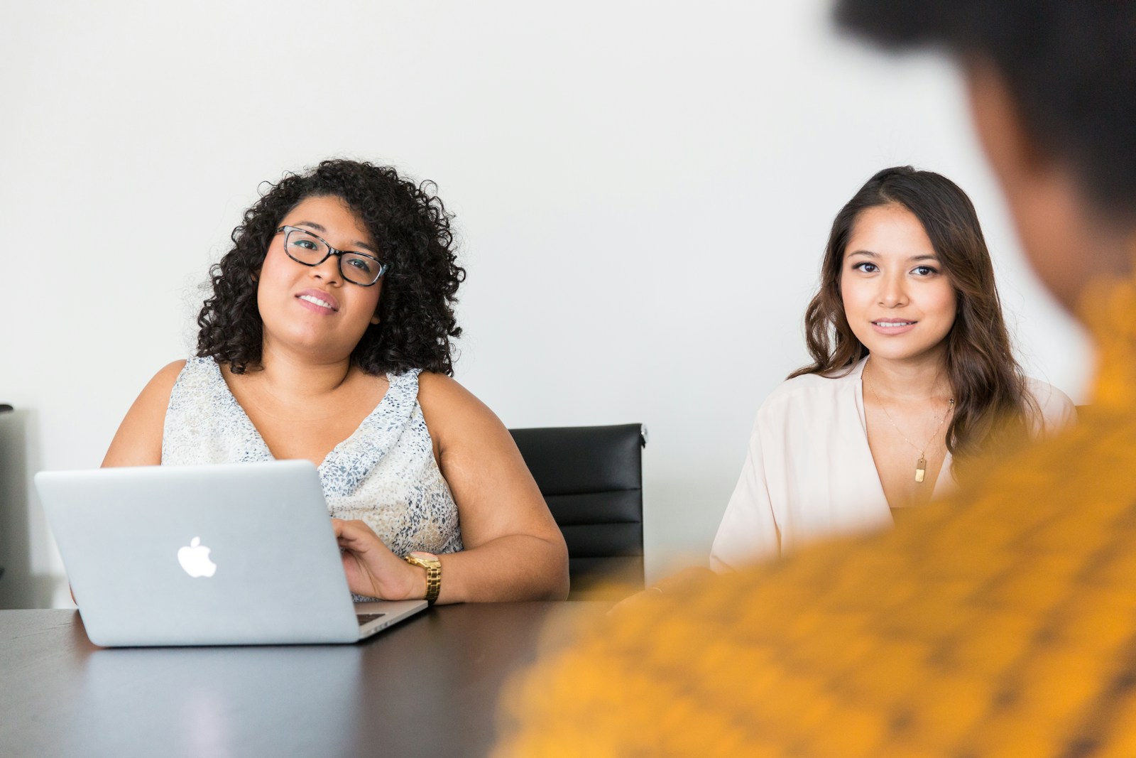 Deux femmes sont assises à une table, l’une d’elles avec un ordinateur portable, face à une troisième personne qui tourne le dos à la caméra, illustrant ainsi des stratégies pour réussir un entretien d’embauche dans un contexte professionnel.