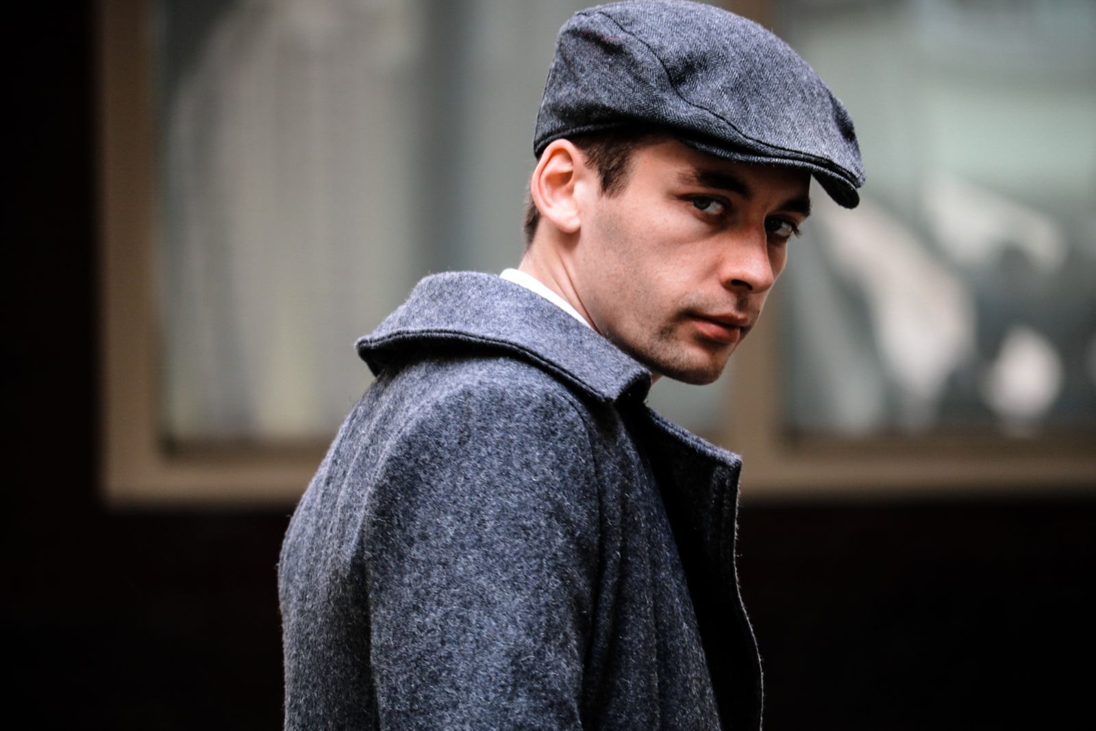 Un homme portant une casquette plate grise et un manteau gris assorti regarde par-dessus son épaule avec un air sérieux, comme s'il réfléchissait à des astuces discrètes pour chercher un emploi, debout devant une fenêtre floue en arrière-plan.