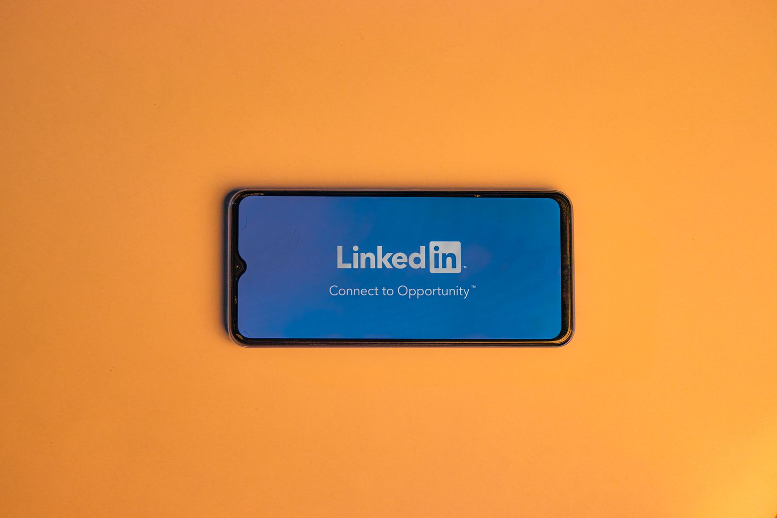 Un smartphone sur lequel l'application LinkedIn est ouverte affiche le message « Connect to Opportunity » sur fond bleu, placé sur une surface orange unie — un rappel motivant pour tous ceux qui cherchent à améliorer leur profil LinkedIn.