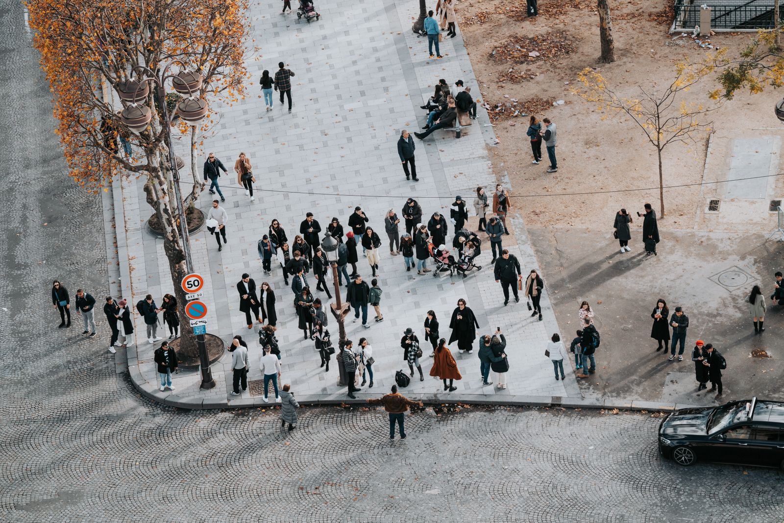 Un groupe de personnes est rassemblé sur un trottoir en ville, près d'un arbre aux feuilles d'automne, dont certaines sont éparpillées aux alentours ; elles prennent peut-être un moment pour activer leur réseau professionnel. Une voiture noire est garée au coin de la rue, et la scène est vue d'en haut.