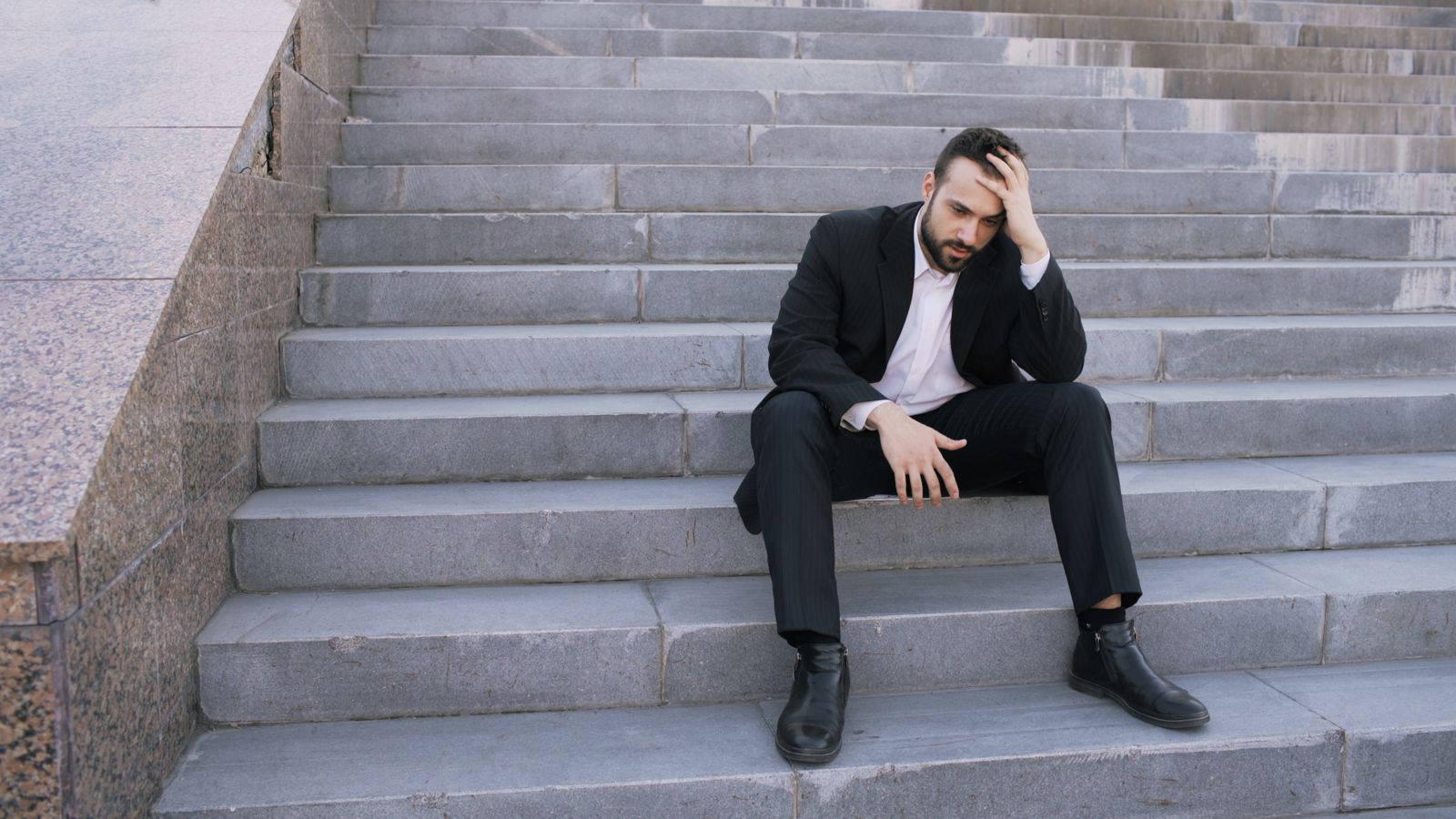 Un homme en costume est assis sur des marches à l'extérieur ; l'air stressé, la tête appuyée sur une main, il s'inquiète de retrouver un emploi après avoir été au chômage.