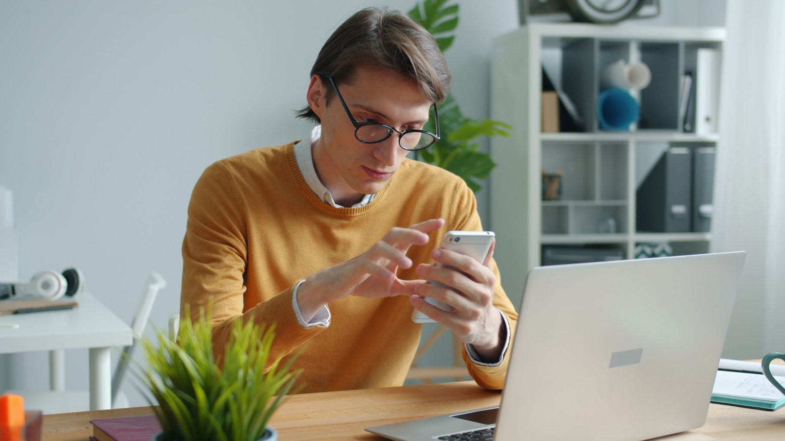Une personne portant des lunettes et un pull couleur moutarde est assise à un bureau devant un ordinateur portable ; elle regarde et utilise son smartphone, peut-être pour relancer une candidature. Des plantes, des livres et des fournitures de bureau occupent l'arrière-plan.