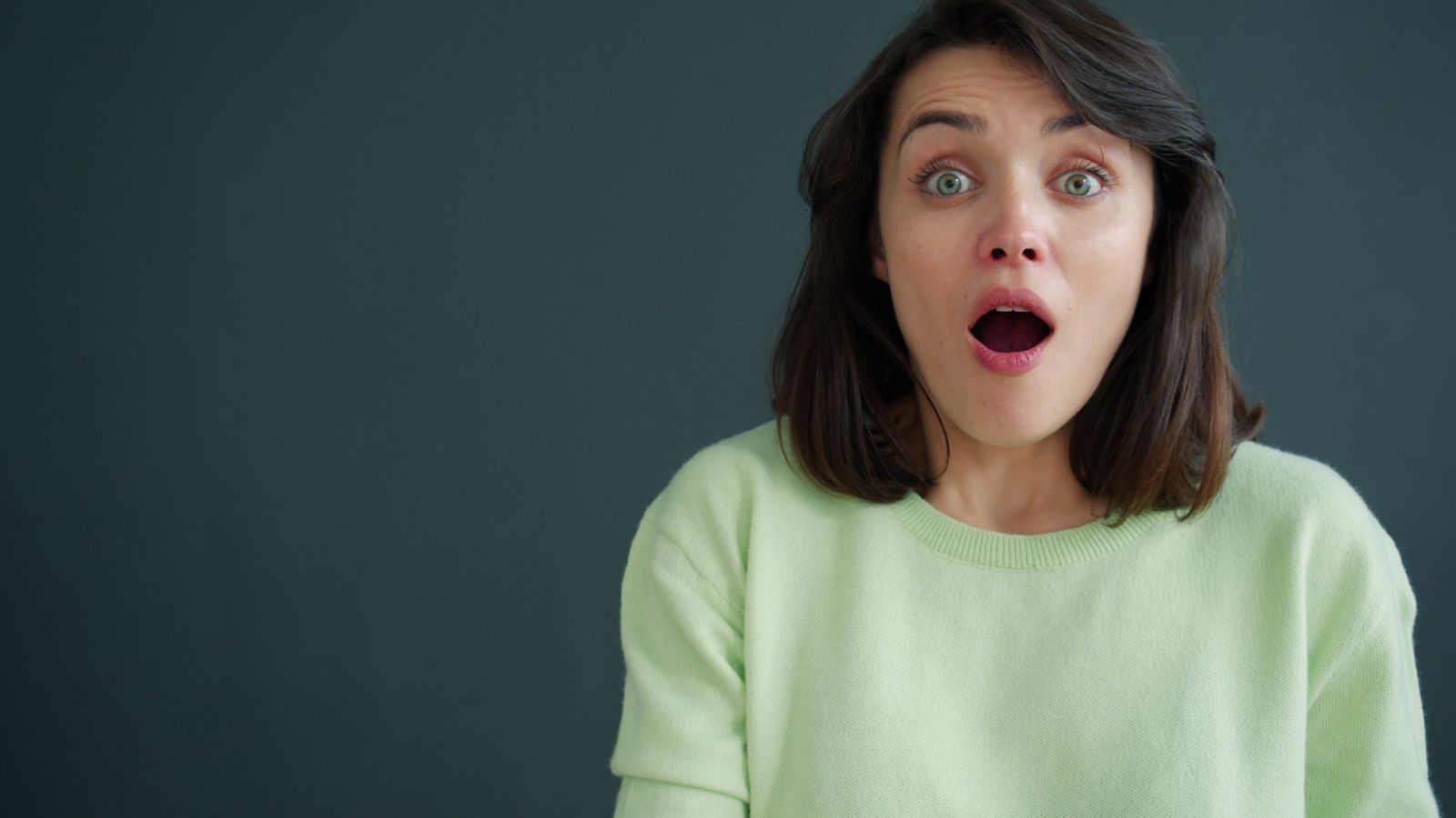 Une femme aux cheveux châtains mi-longs, vêtue d'un pull vert clair, a l'air surprise, les yeux écarquillés et la bouche ouverte, comme si elle réagissait à des questions pièges posées lors d'un entretien, sur un fond sombre et uni.