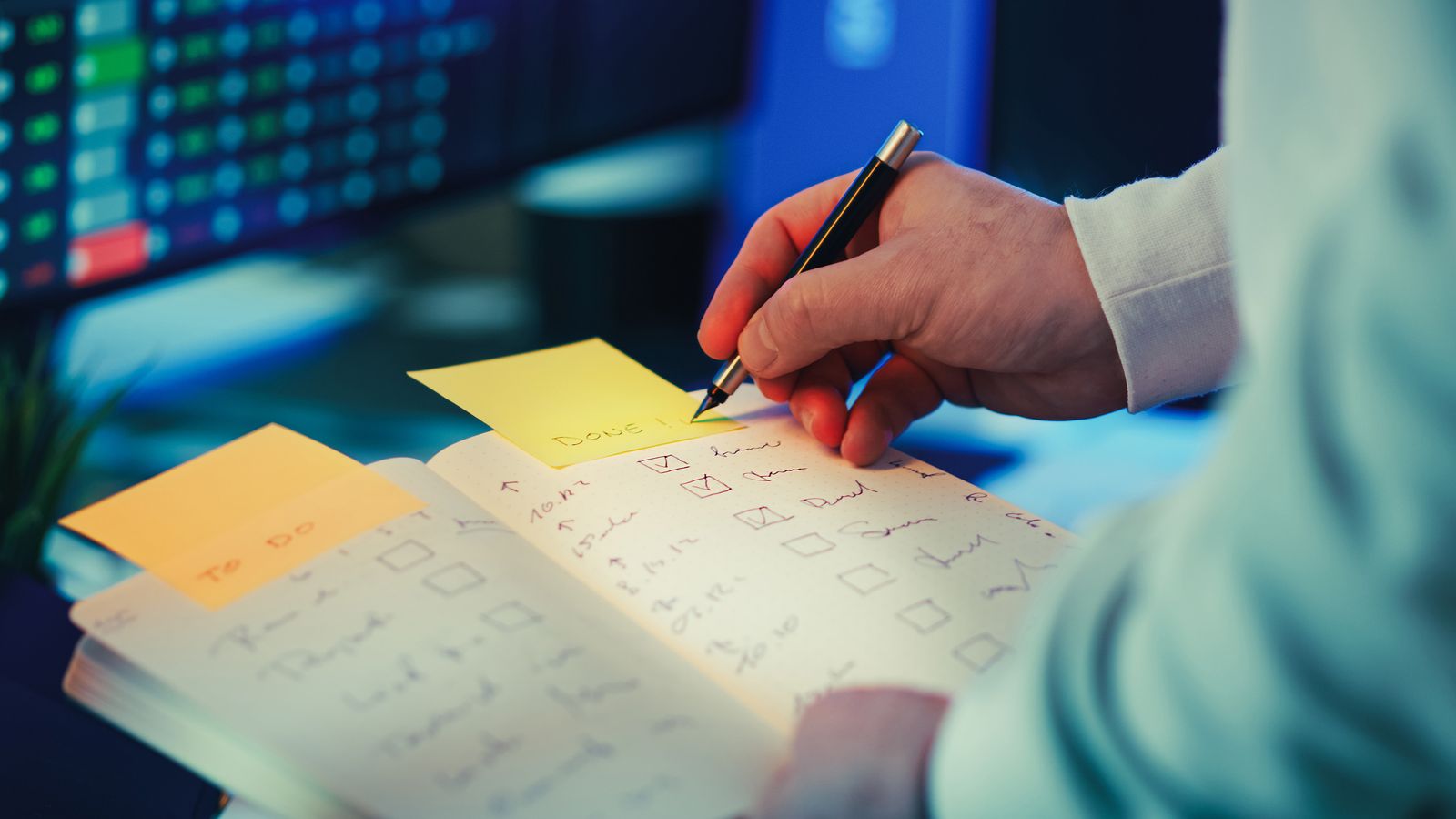 Une personne écrit sur un post-it avec un stylo tout en tenant un cahier ouvert sur lequel figurent des listes de tâches manuscrites accompagnées de cases à cocher — illustrant ainsi des astuces pour organiser sa recherche d'emploi dans un espace de travail bien éclairé et propice à la productivité.