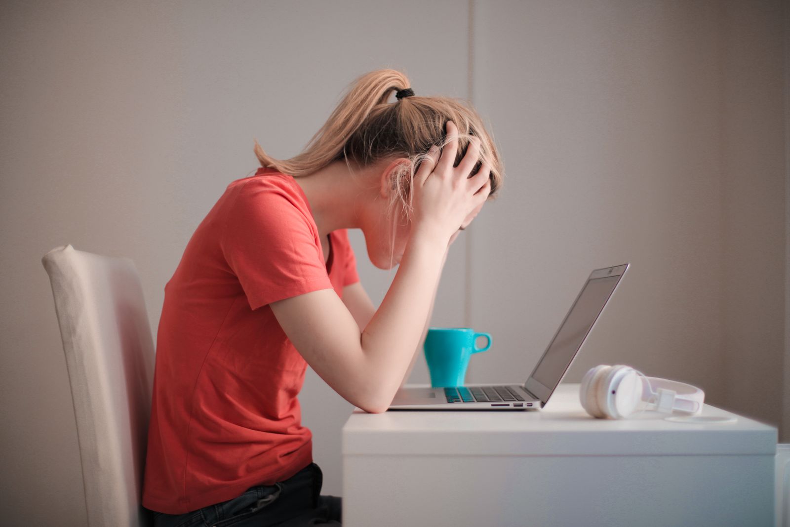 Une femme vêtue d'un t-shirt rouge est assise à un bureau, la tête entre les mains, l'air stressée, face à un ordinateur portable ouvert — peut-être après avoir rencontré des erreurs qui font échouer une candidature spontanée. Une tasse bleue et un casque audio blanc se trouvent à côté d'elle.