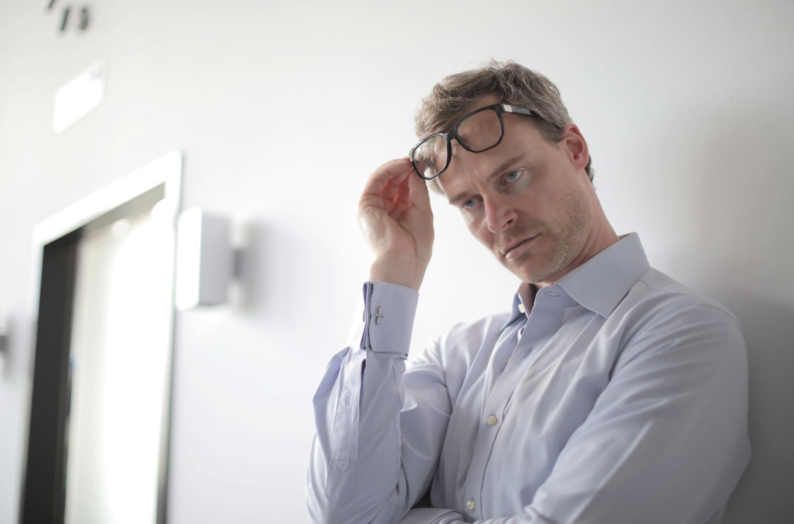 Un homme vêtu d'une chemise bleu clair se tient debout devant un mur blanc, tenant ses lunettes d'une main et affichant un air pensif ou inquiet, peut-être en train de réfléchir aux erreurs commises dans son parcours professionnel.