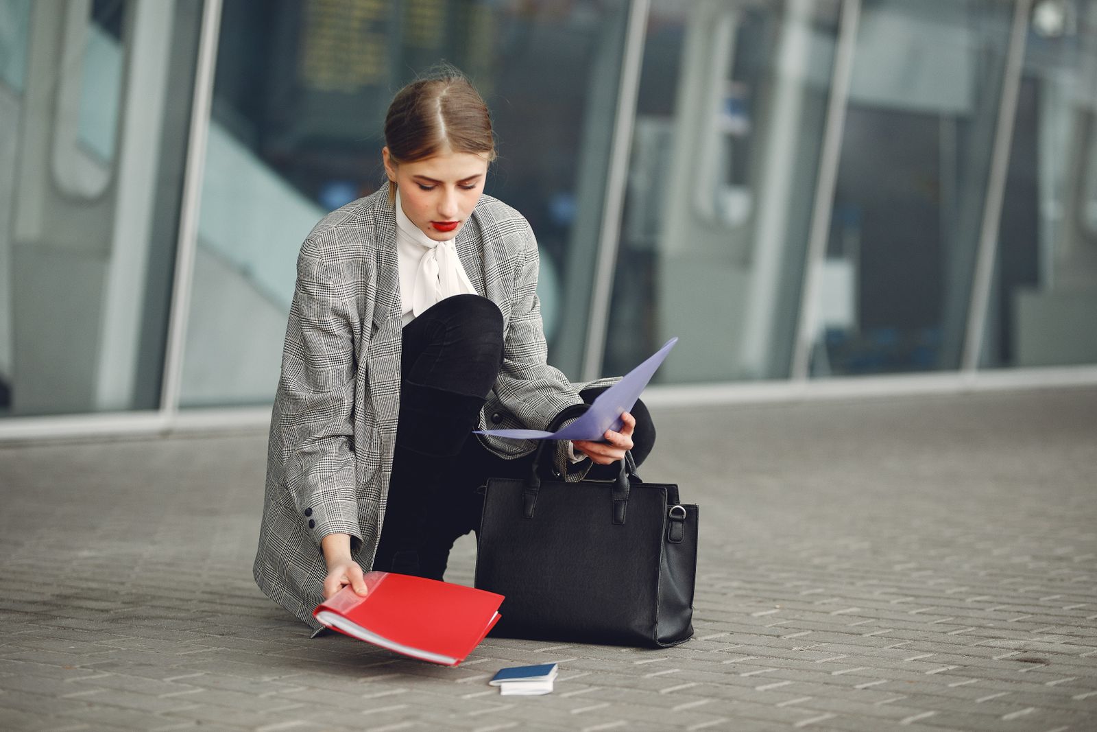 Une femme en tenue professionnelle est agenouillée sur le trottoir, ramassant des dossiers et des papiers éparpillés à côté d'un sac à main noir, devant un immeuble aux façades vitrées — illustrant peut-être la difficulté à laquelle sont confrontés nombre de ceux qui tentent de trouver un emploi sans diplôme.