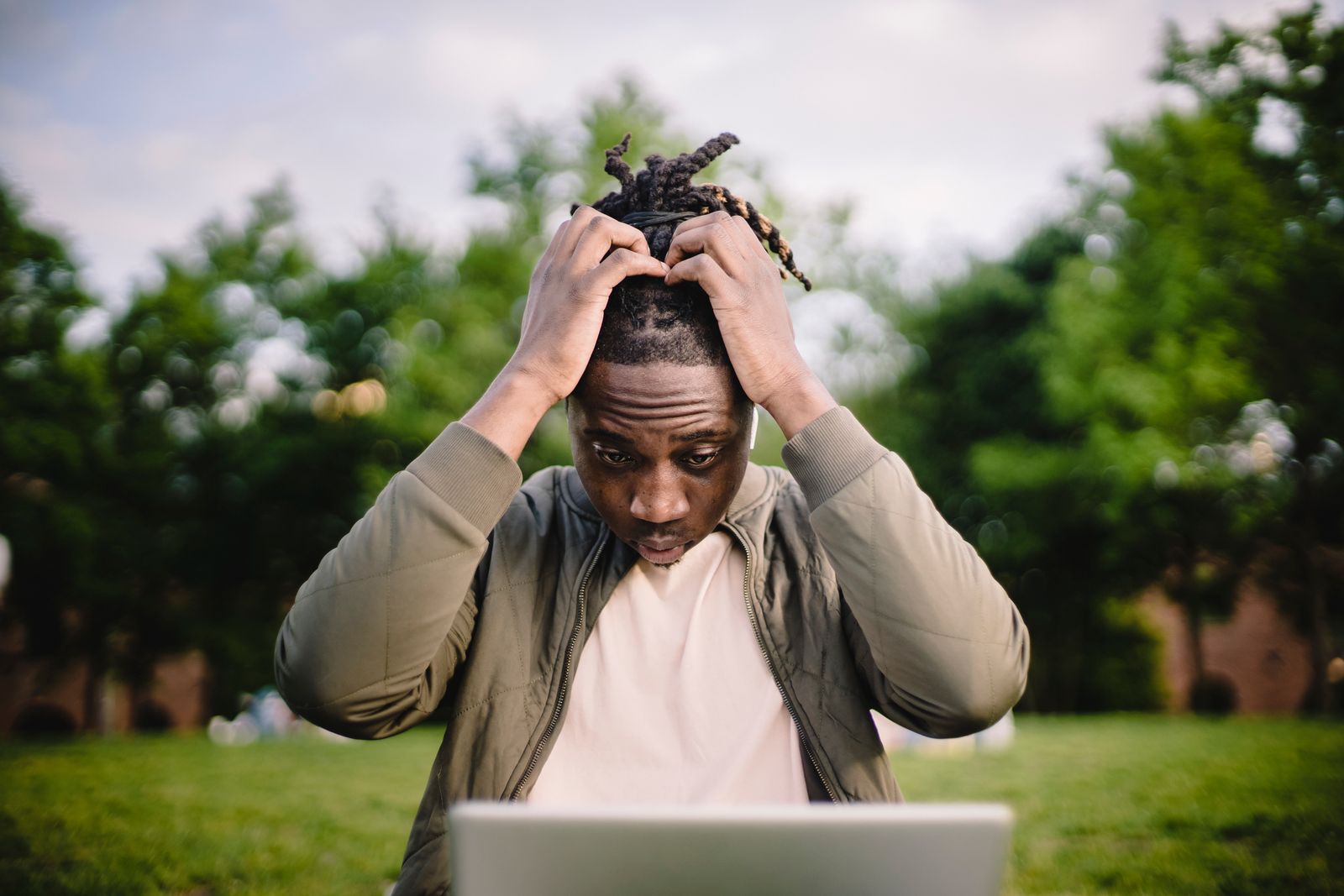 Une personne assise dehors sur l'herbe, l'air stressé ou frustré, se tenant la tête à deux mains et fixant un ordinateur portable — peut-être inquiète à cause d'erreurs dans un e-mail de candidature. On aperçoit des arbres et de la verdure en arrière-plan.