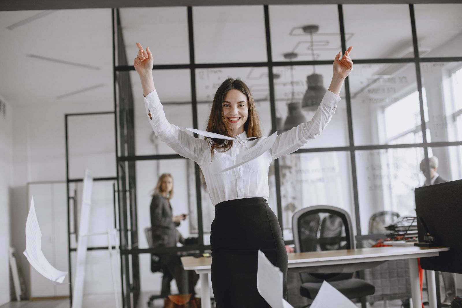 Une femme en tenue professionnelle se tient debout dans un bureau ; elle sourit et lance des papiers en l'air, célébrant ainsi les conseils pour sa reconversion réussie. Une autre personne travaille à un bureau près de grandes baies vitrées.