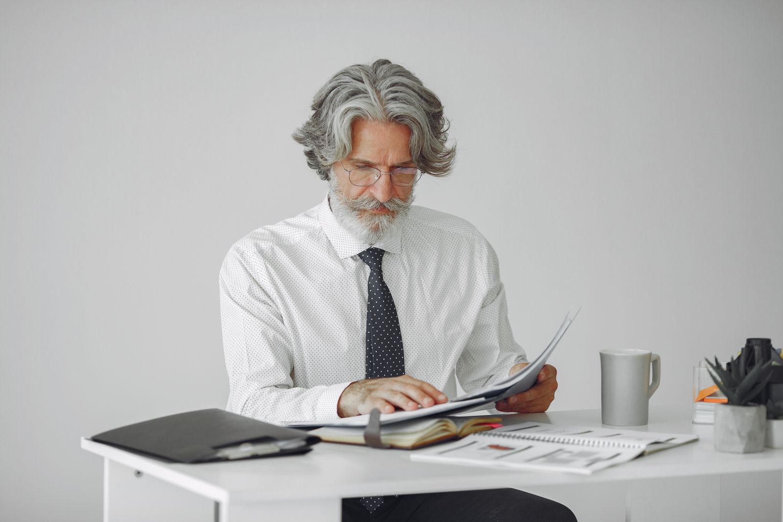 Un homme d'un certain âge, aux cheveux gris et à la barbe, portant des lunettes et une cravate, est assis à un bureau en train de lire des documents. Entouré de papiers, d'une tasse et de fournitures de bureau posés sur le bureau blanc, il semble examiner attentivement des « conseils pour les candidats seniors ».