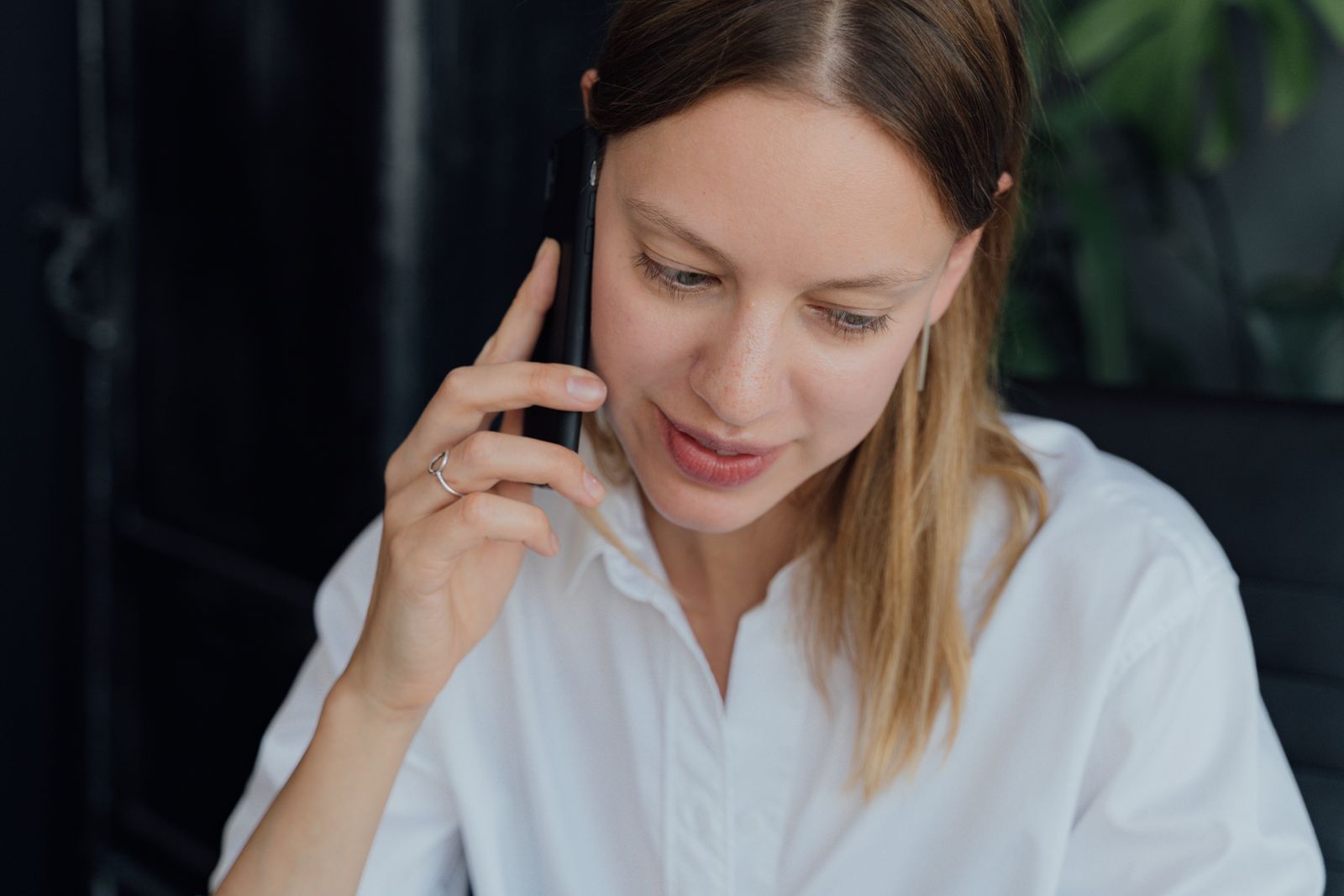 Une femme aux cheveux raides châtain clair, vêtue d'une chemise blanche, est en train de parler au téléphone. Elle a le regard baissé et semble engagée dans une conversation sérieuse, peut-être à la recherche de conseils pour relancer un recruteur.