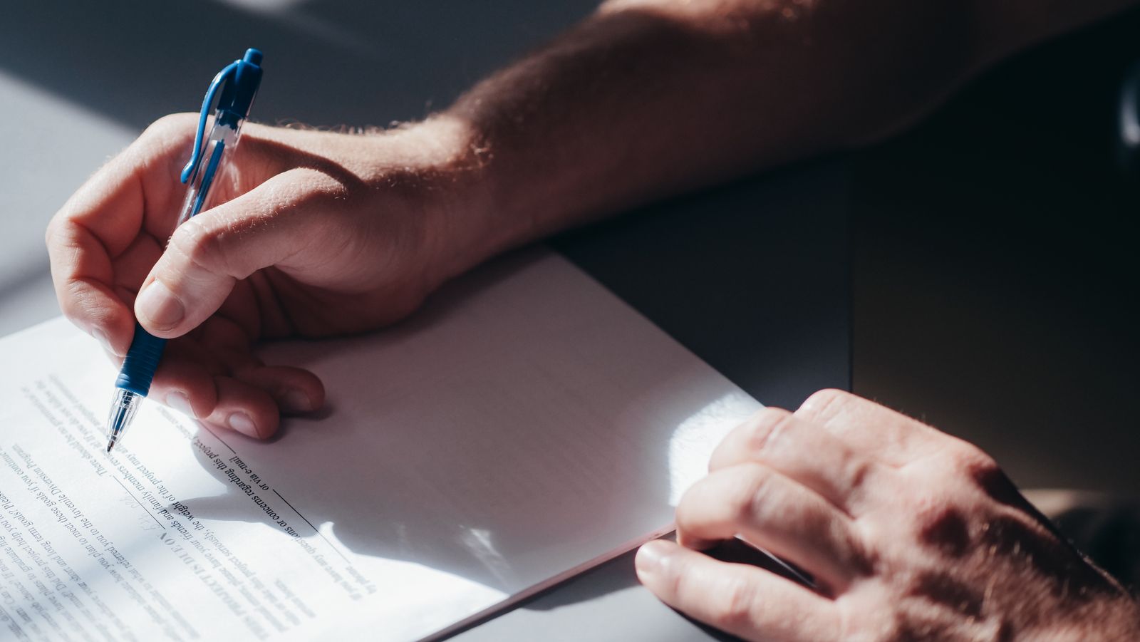 Une personne tenant un stylo bleu s'apprête à signer ou à écrire sur un document posé sur un bureau, vérifiant peut-être une dernière fois s'il y a des erreurs dans la lettre de motivation, tandis que la lumière du soleil projette des ombres sur le papier.