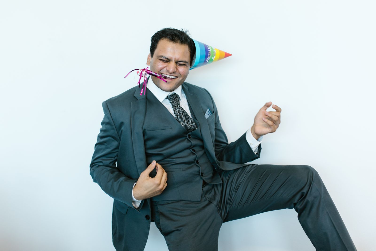 Un homme en costume, coiffé d'un chapeau de fête coloré, souffle dans une trompette de fête et prend la pose de manière espiègle devant un fond blanc, célébrant comme s'il venait de réussir sa reconversion professionnelle.
