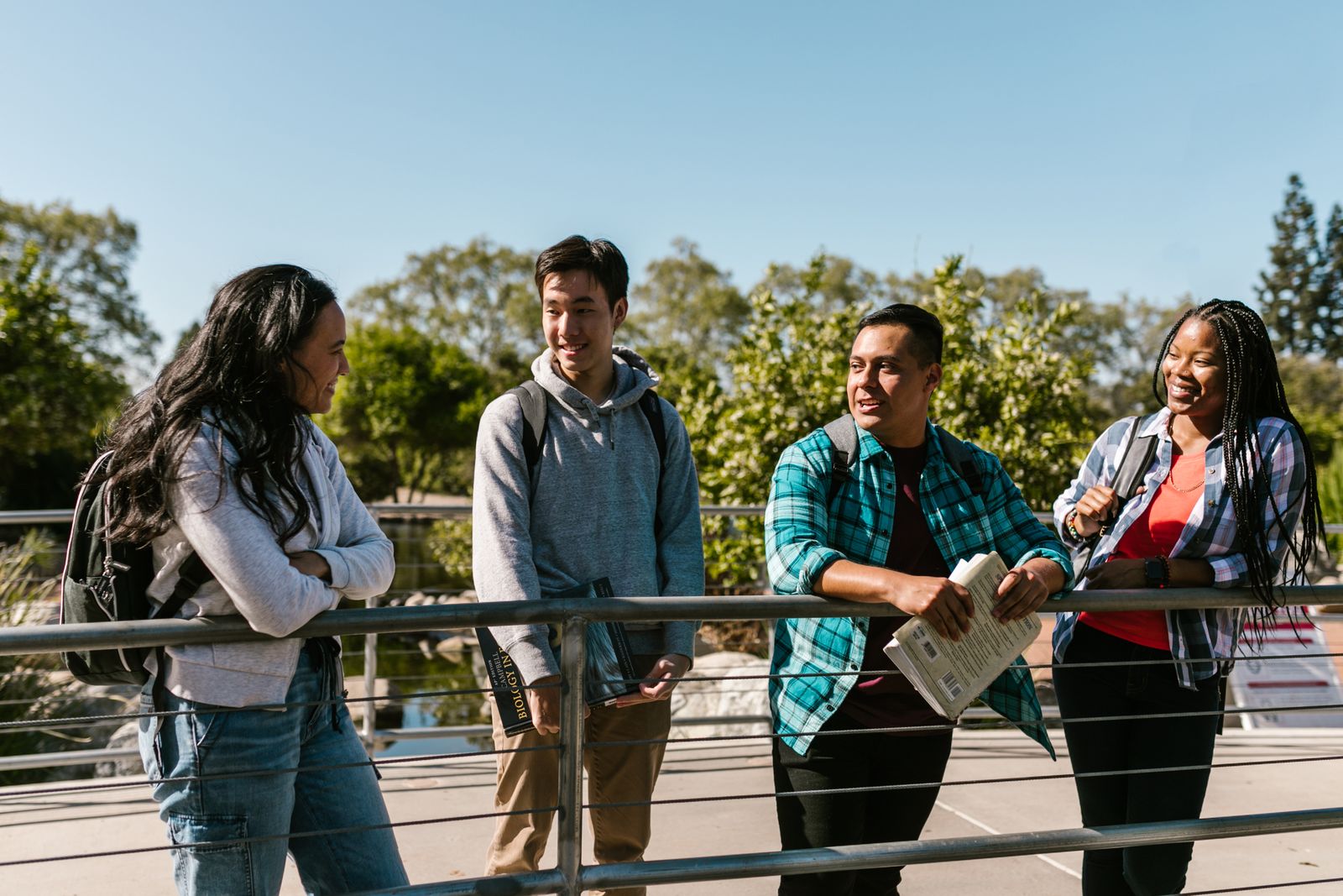 15 conseils oubliés pour trouver un stage ou une alternance plus vite Quatre jeunes adultes se tiennent à l'extérieur sur un pont, souriants et en pleine conversation. Vêtus de manière décontractée, ils semblent échanger des conseils pour trouver un stage par une journée ensoleillée, avec des arbres et un ciel bleu en arrière-plan.