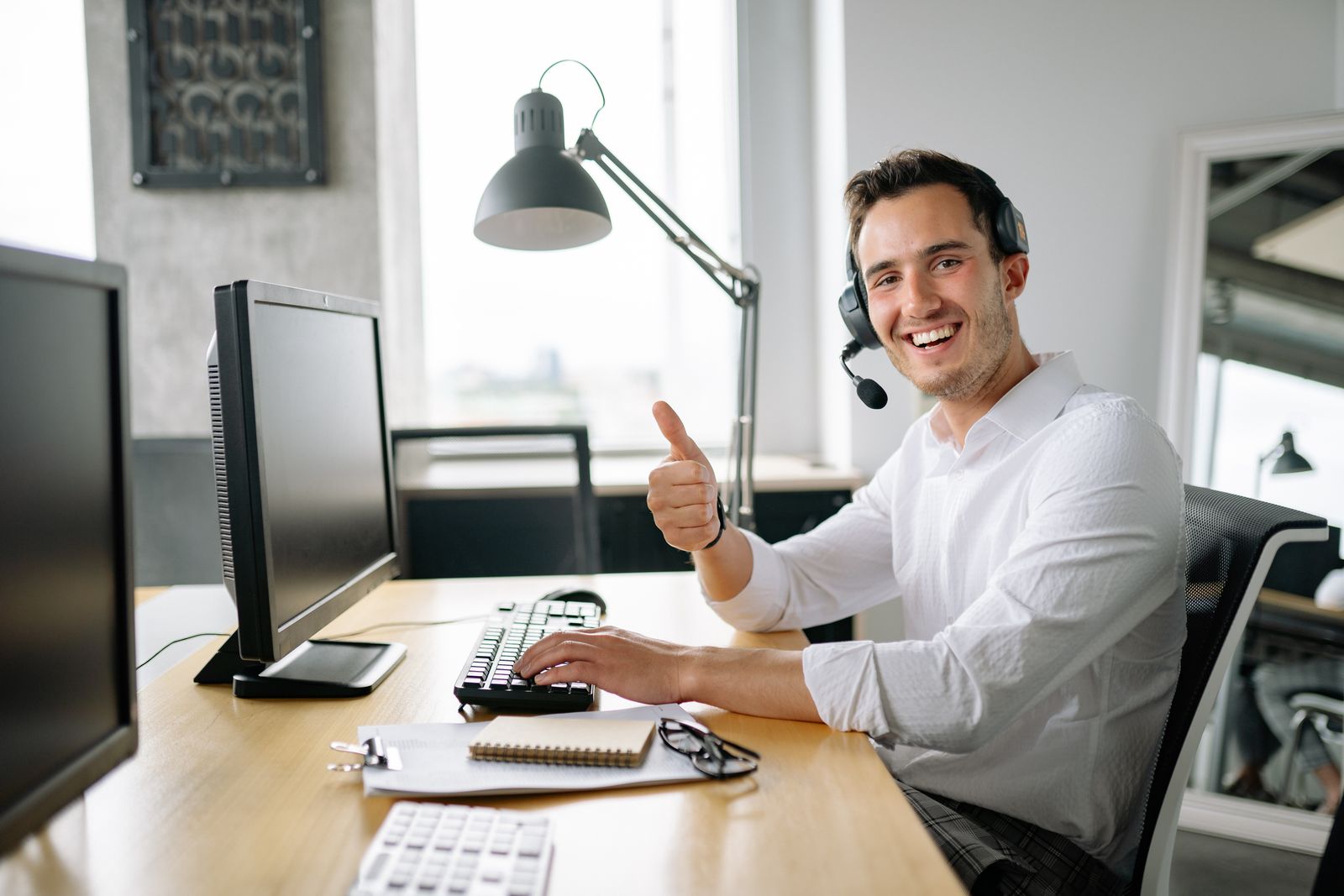Un homme souriant, équipé d'un casque, est assis devant un bureau équipé d'un ordinateur et lève le pouce en signe d'approbation. Dans son bureau moderne baigné de lumière naturelle, il cherche à trouver les bonnes offres d'emploi, un bloc-notes et un clavier posés devant lui.