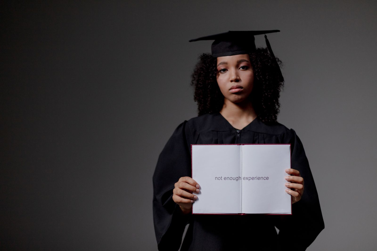 Une diplômée, vêtue d'une toge et d'un mortier, tient un livre ouvert sur lequel on peut lire « pas assez d'expérience », le visage impassible, devant un fond uni, symbolisant les difficultés rencontrées par de nombreuses personnes à la recherche de conseils pour trouver un emploi sans expérience.